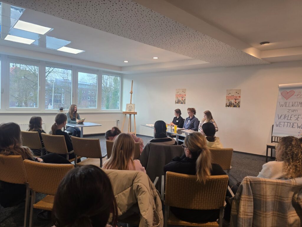 Schülerin, die links sitzend vorliest. Jury auf der rechten Seite. Publikum zuhörend im Vordergrund. 